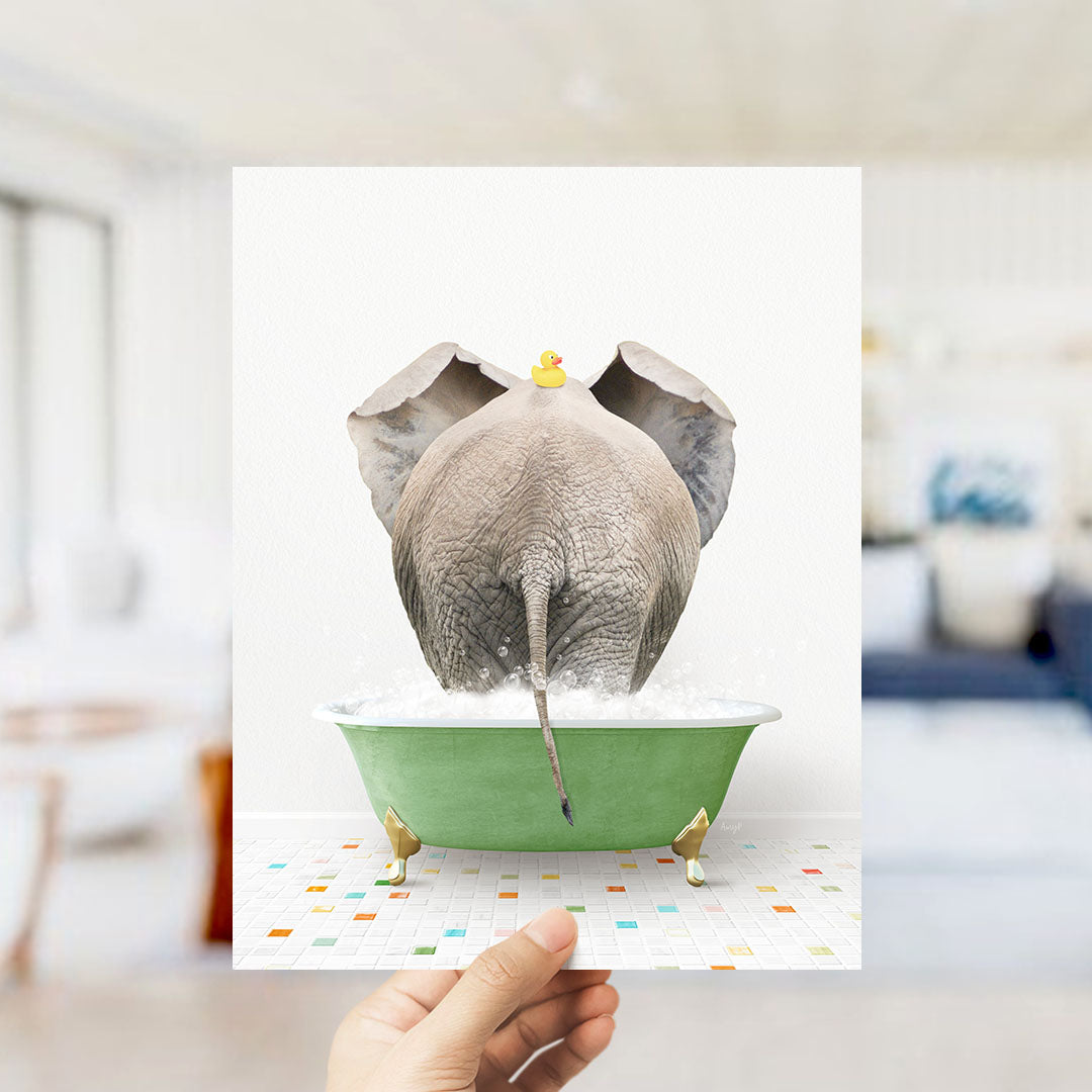 A hand holds up a framed image depicting an elephant sitting in a green bathtub filled with water, with a yellow rubber duck floating on its head.