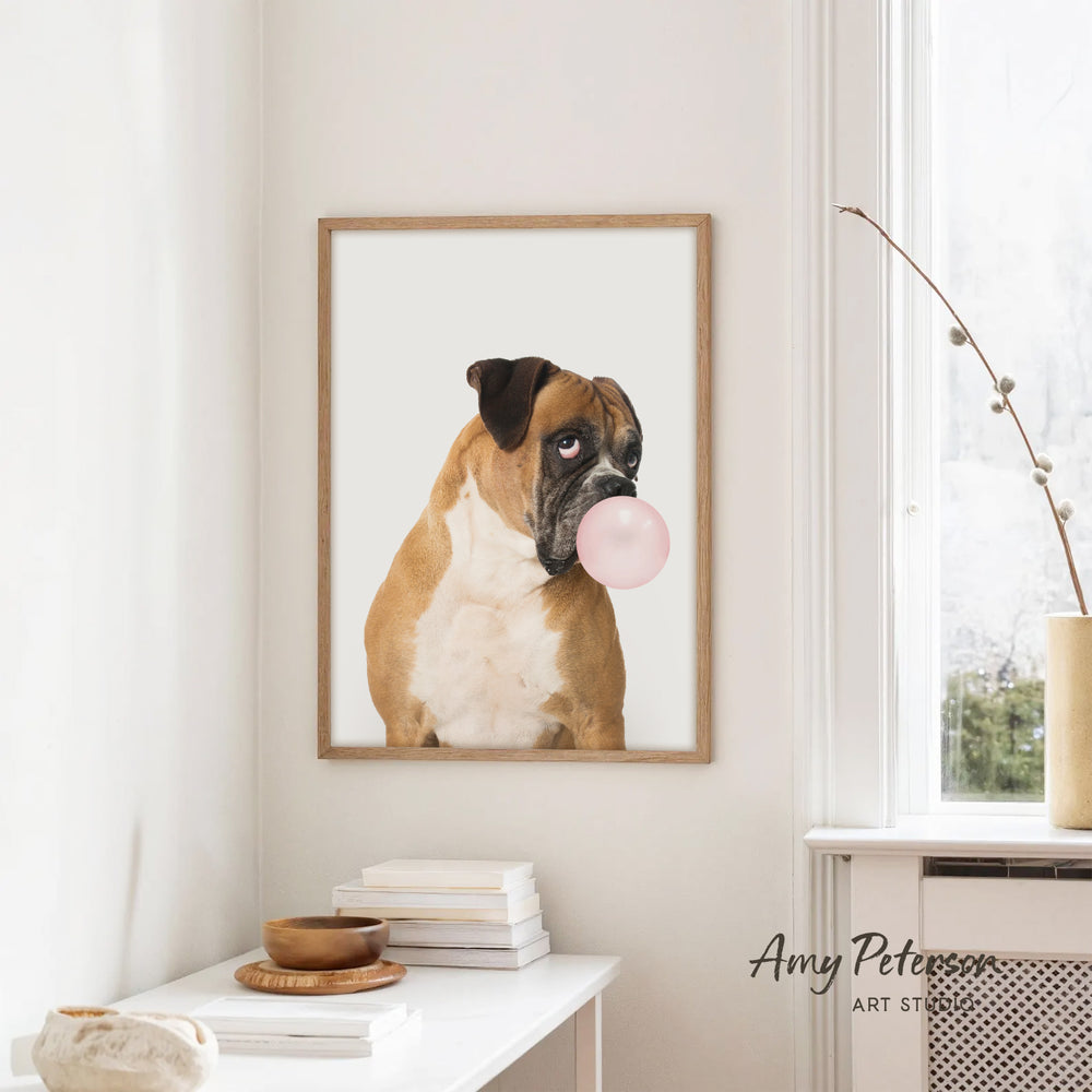A framed photograph of a brown and white dog blowing a bubble gum bubble is hanging on a white wall in a room with a white table and a window.