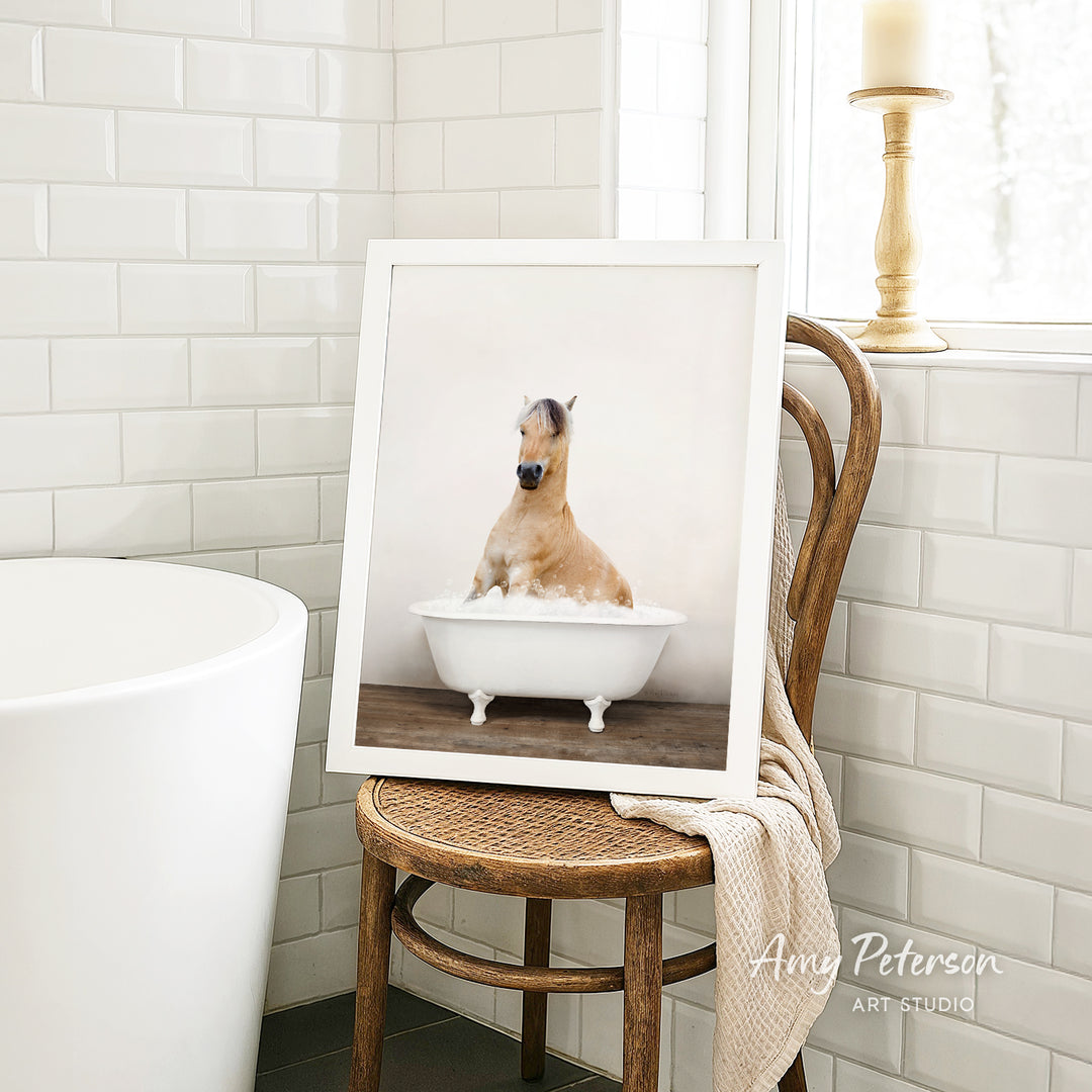A framed photograph of a horse sitting in a bathtub, with a towel draped over the back of a wooden chair in a bathroom setting.