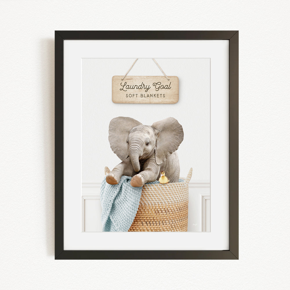 A framed image of a baby elephant sitting in a woven basket, with a sign above it that reads "Laundry Coal Soft Blankets".