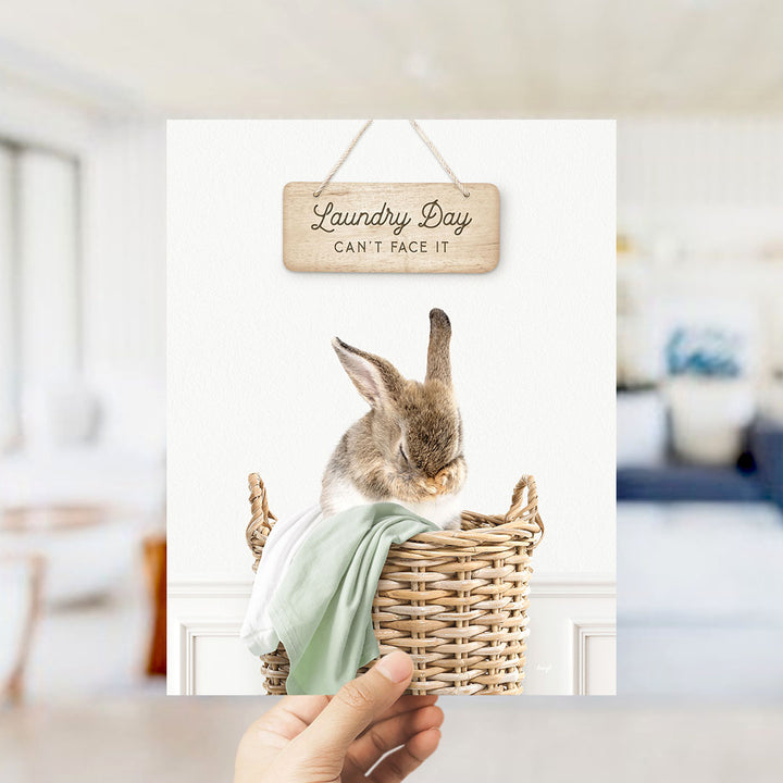A hand holding a wooden sign that says "Laundry Day Can't Face It" above a basket containing a small rabbit.