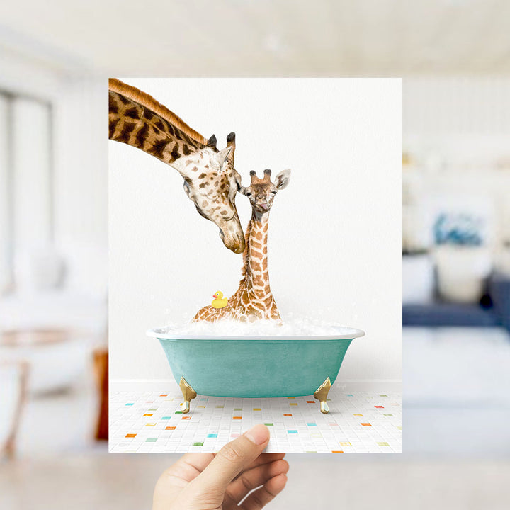 A hand holds up a framed image depicting two giraffes in a bathtub, with one giraffe gently nuzzling the other.