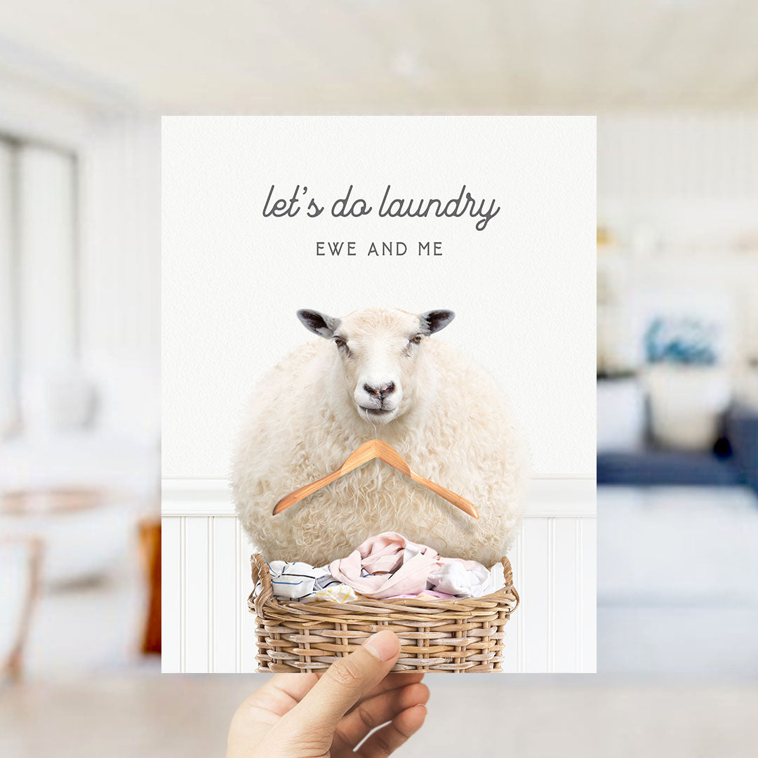 A hand holding a card with a sheep sitting in a basket of laundry, with the text "let's do laundry EWE AND ME" written on it.