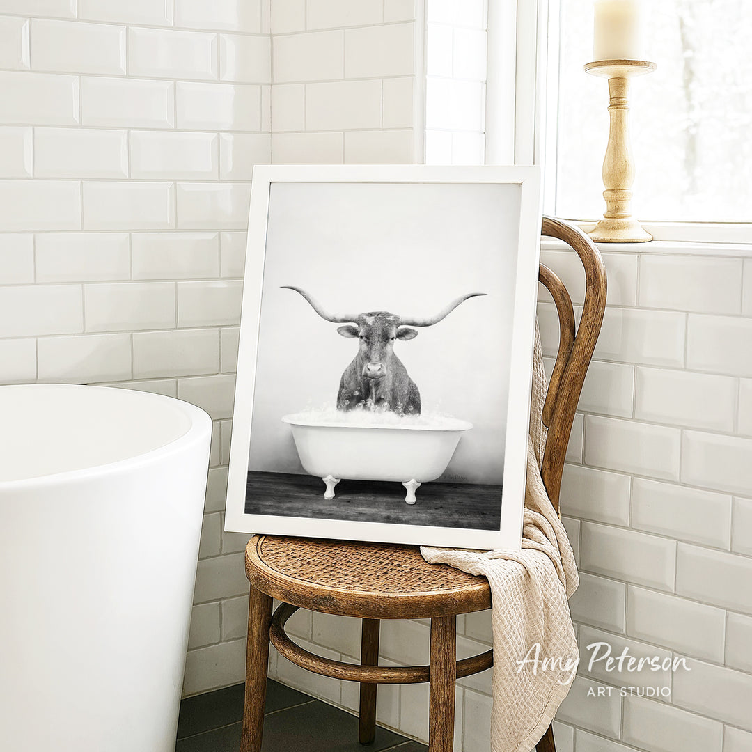 A framed black and white photograph of a bull with long horns sitting in a bathtub is displayed on a wooden chair in a bathroom.