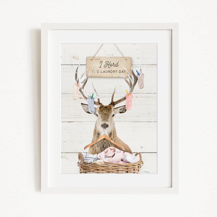 A framed image depicting a deer with antlers holding laundry, accompanied by a sign that reads "I herd it's laundry day".