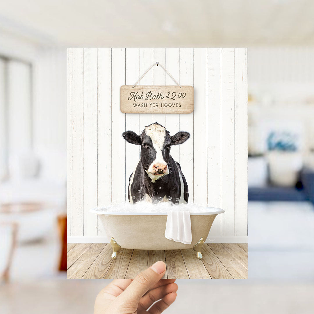 A hand holding a framed image of a black and white cow sitting in a bathtub, with a sign above it advertising hot baths for $2.00.