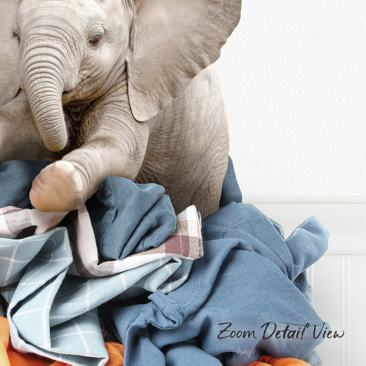 A baby elephant sitting on a pile of clothes, with its trunk curled up.