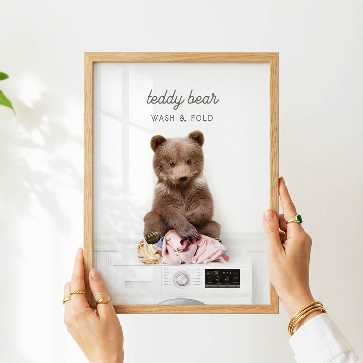 A person's hands holding a framed image of a teddy bear sitting on a washing machine, with the text "teddy bear WASH & FOLD" displayed on the frame.