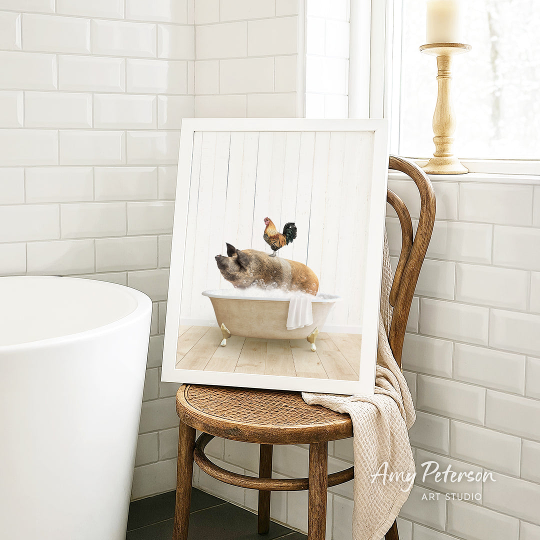 A framed image of a cat sitting in a bathtub, with a rooster perched on its head, is displayed on a wooden chair in a bathroom setting.