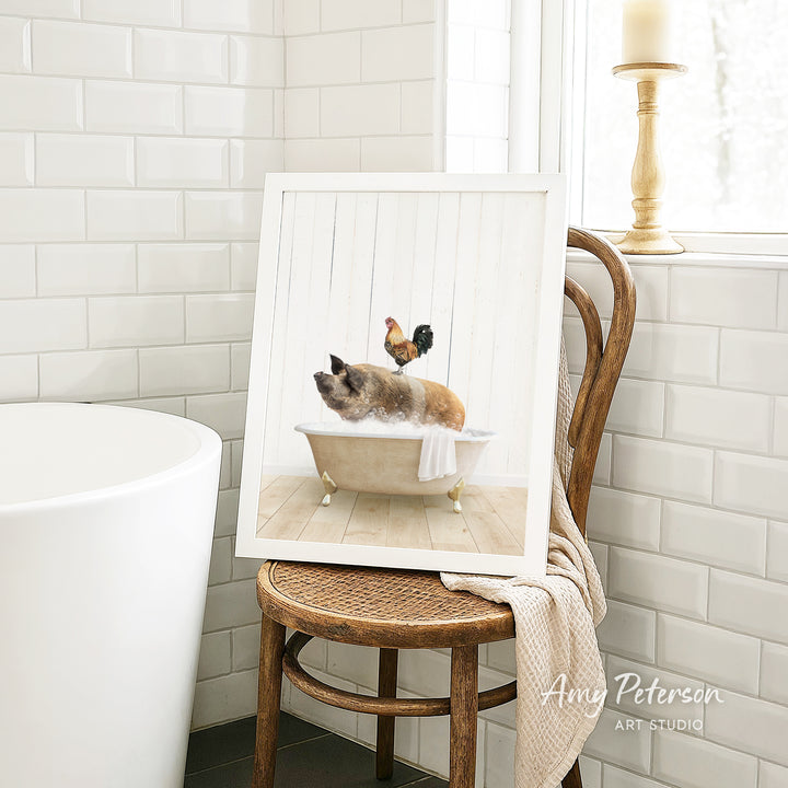 A framed image of a cat sitting in a bathtub, with a rooster perched on its head, is displayed on a wooden chair in a bathroom setting.