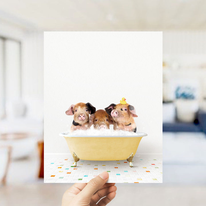 A hand holds up a framed image of three adorable pigs sitting in a yellow bathtub filled with bubbles, with a yellow rubber duck perched on the side.