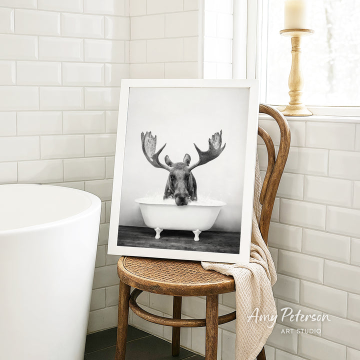 A framed black and white photograph of a moose sitting in a bathtub is displayed on a wooden chair in a bathroom setting.