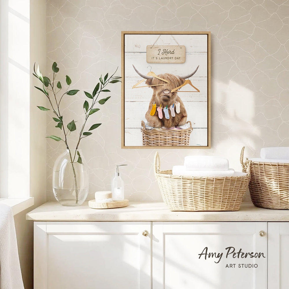 a framed picture of a Highland cow hanging out of a laundry basket, with a sign above it that says "I herd it's laundry day". The picture is displayed on a wall above a white cabinet, and there is a vase with a plant and a soap dispenser on the cabinet.