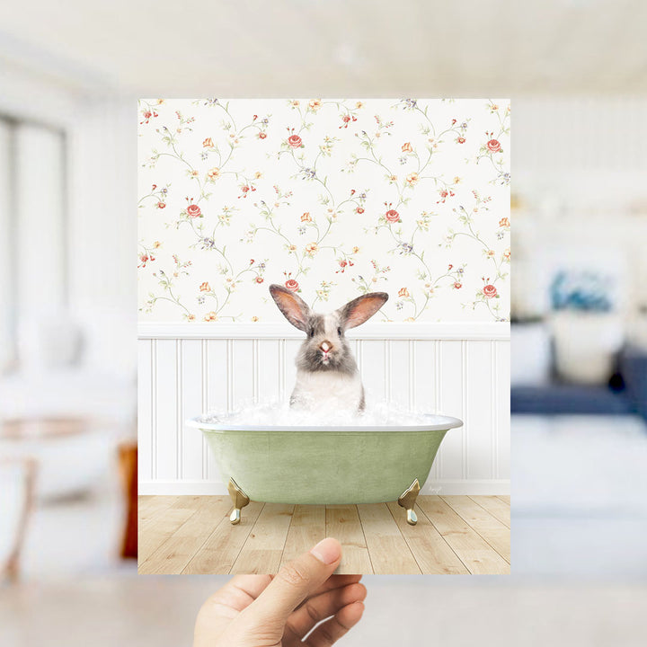 A hand holds up a framed image of a rabbit sitting in a green bathtub filled with water, with a floral wallpaper in the background.