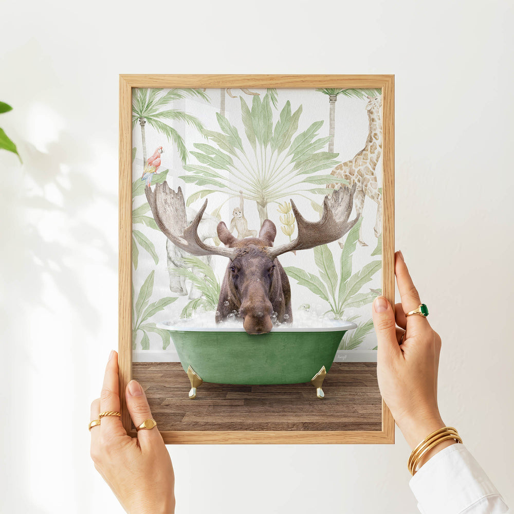 A person's hands are holding a framed picture of a moose in a bathtub, surrounded by tropical foliage.