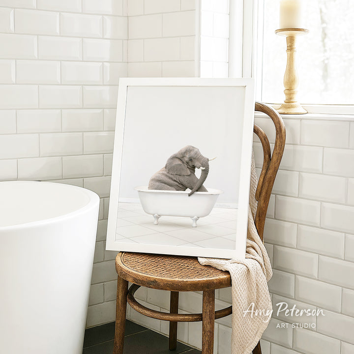 A framed image of a cat sitting in a bathtub is displayed on a wooden chair in a bathroom setting.
