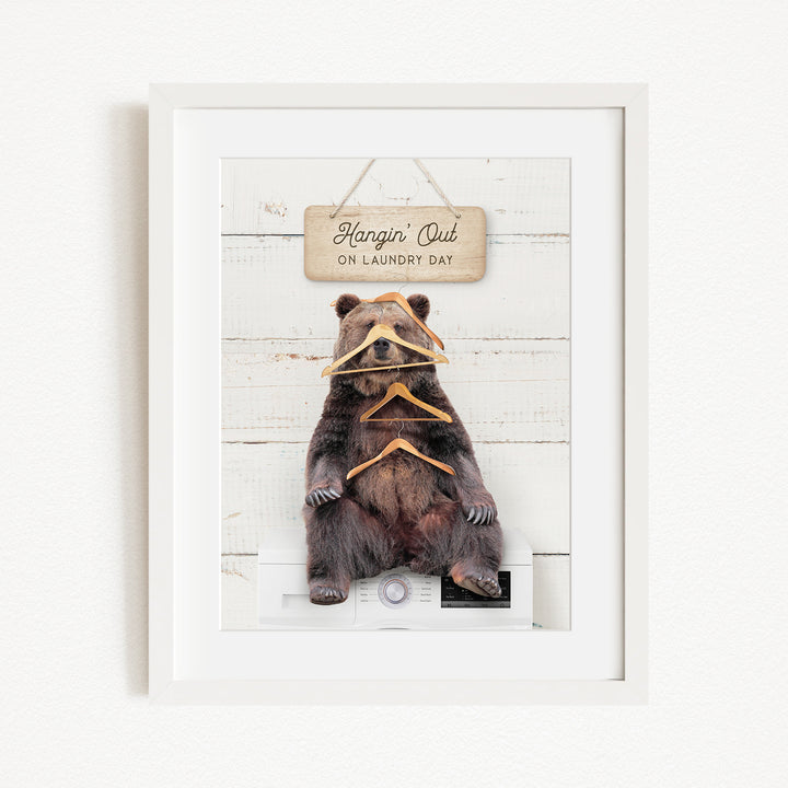A framed image of a brown bear sitting on a washing machine, with a sign above it that says "Hangin' Out on laundry day".
