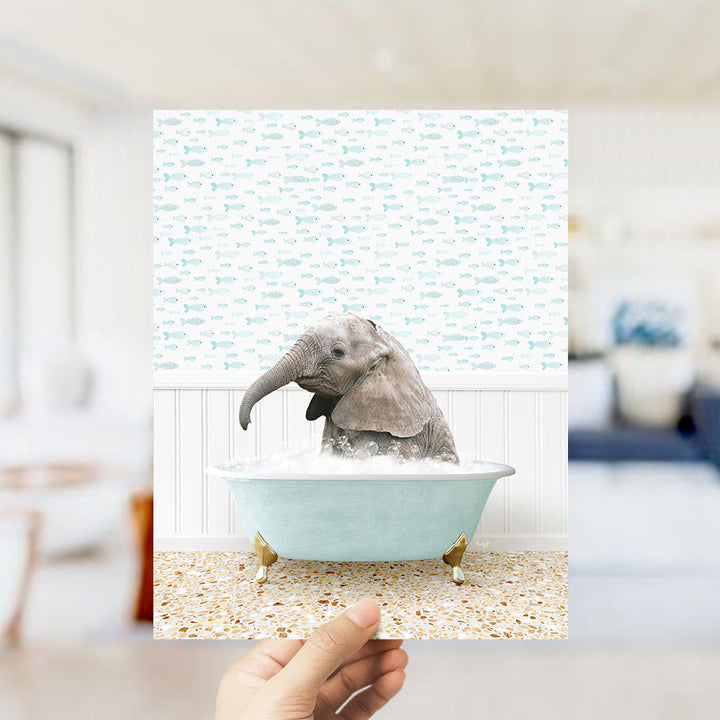 A hand holds up a framed image depicting a baby elephant sitting in a bathtub filled with bubbles, surrounded by a pattern of small fish.