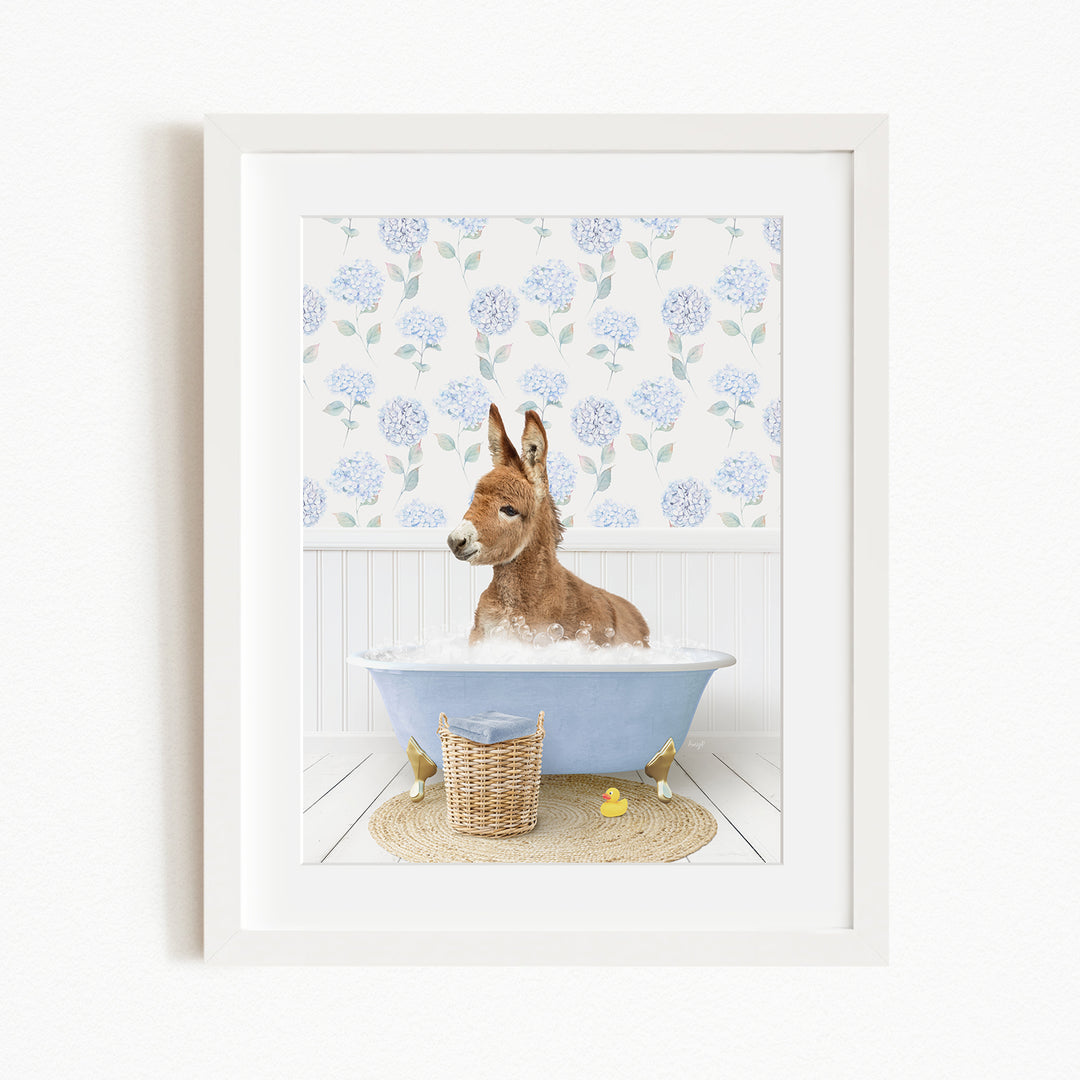 A donkey is sitting in a blue bathtub filled with bubbles, surrounded by a floral patterned wall and a basket of rubber ducks.
