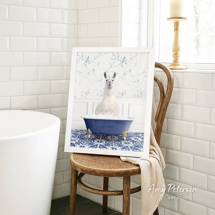 A framed image of a white llama sitting in a blue bathtub, surrounded by a blue and white floral pattern, is displayed on a wooden chair in a bathroom setting.