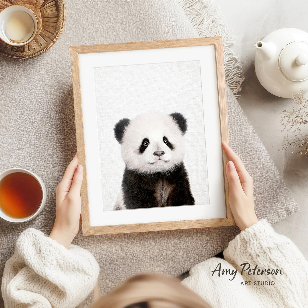 A person is holding a framed picture of a panda bear, which is the main focus of the image. The panda bear is sitting on a white surface, and there is a teapot and a cup nearby. The background of the image is a light gray color.