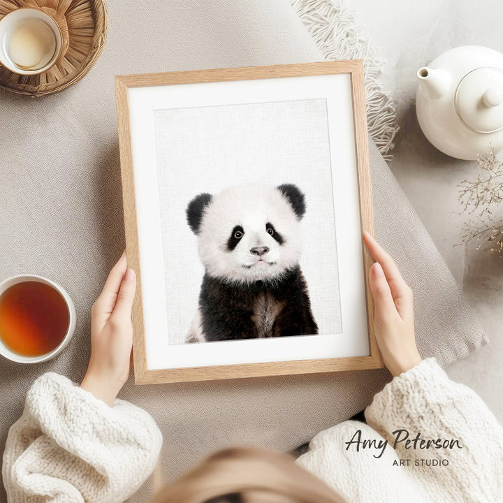 A person is holding a framed picture of a panda bear, which is the main focus of the image. The panda bear is sitting on a white surface, and there is a teapot and a cup nearby. The background of the image is a light gray color.