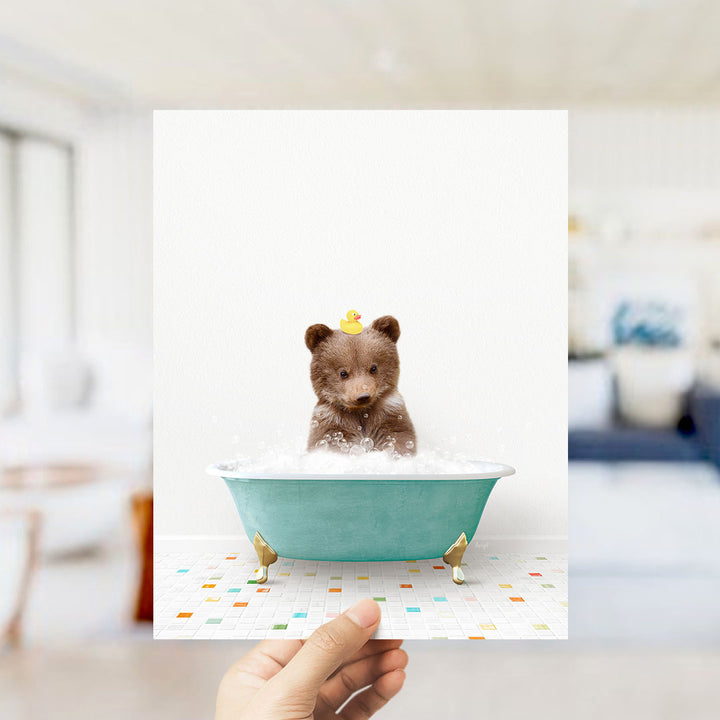 A hand holds up a framed image of a small brown bear sitting in a blue bathtub filled with bubbles, with a yellow rubber duck perched on its head.