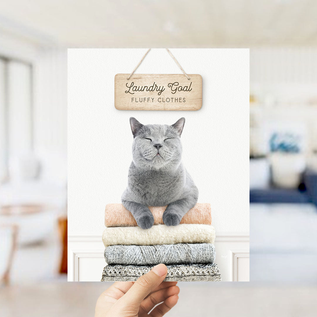 A hand is holding up a framed image of a gray cat sitting on a stack of clothes, with a wooden sign above it that says "Laundry Goal Fluffy Clothes".