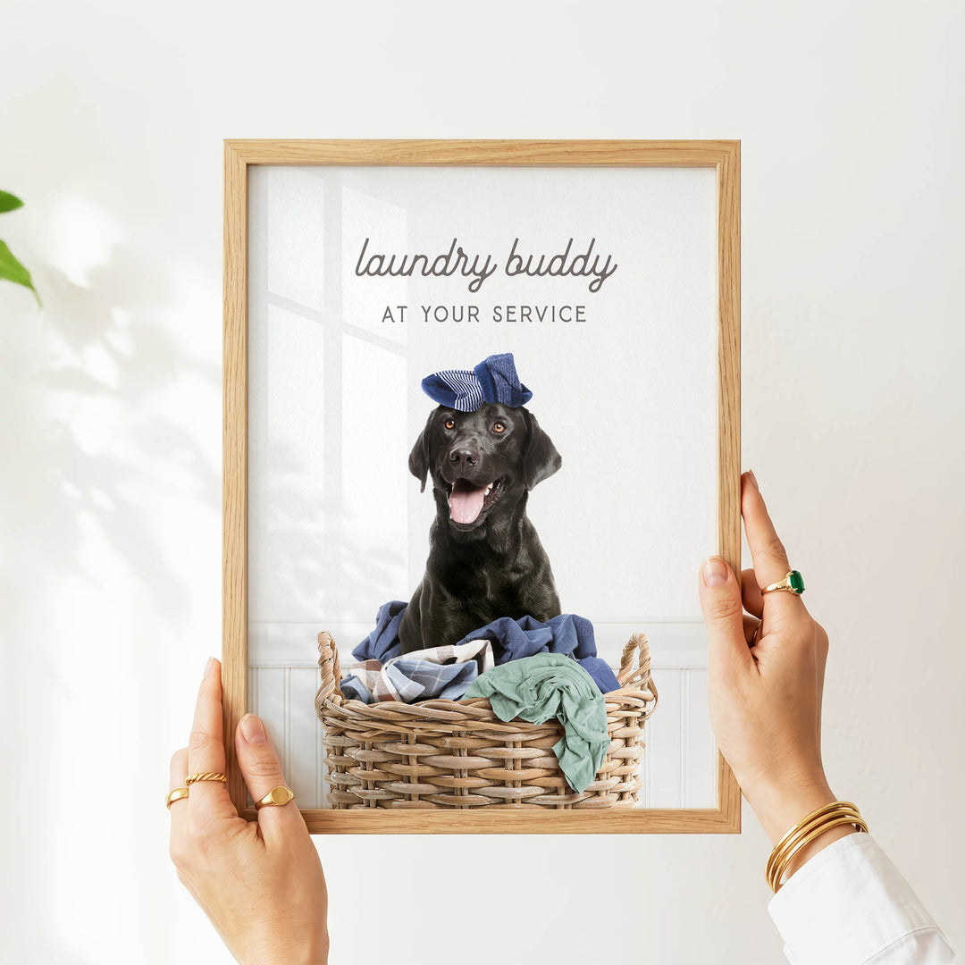 A person is holding up a framed photo of a black dog sitting in a wicker basket filled with clothes, with the text "laundry buddy at your service" written above the image.