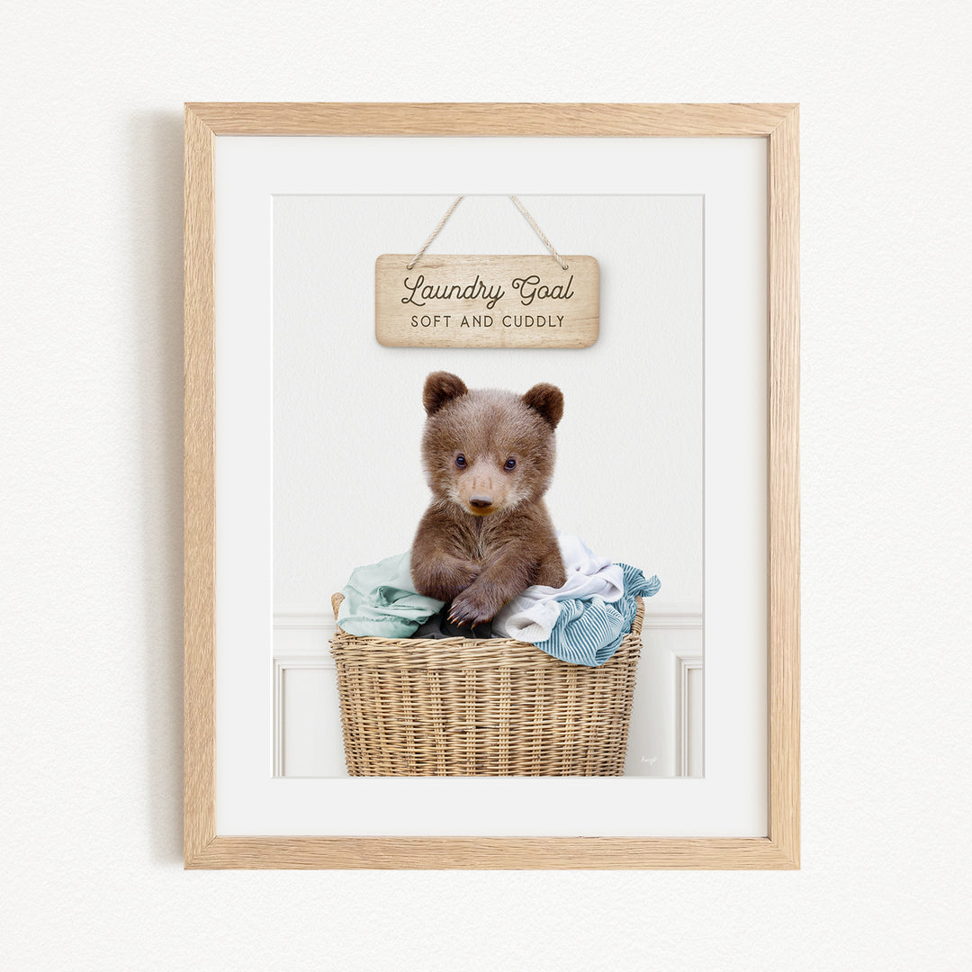 A framed photo of a baby bear sitting in a wicker laundry basket, with a wooden sign above it that says "Laundry Goal Soft and Cuddly".