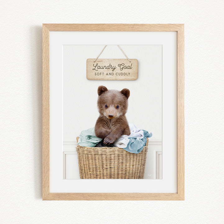 A framed photo of a baby bear sitting in a wicker laundry basket, with a wooden sign above it that says "Laundry Goal Soft and Cuddly".