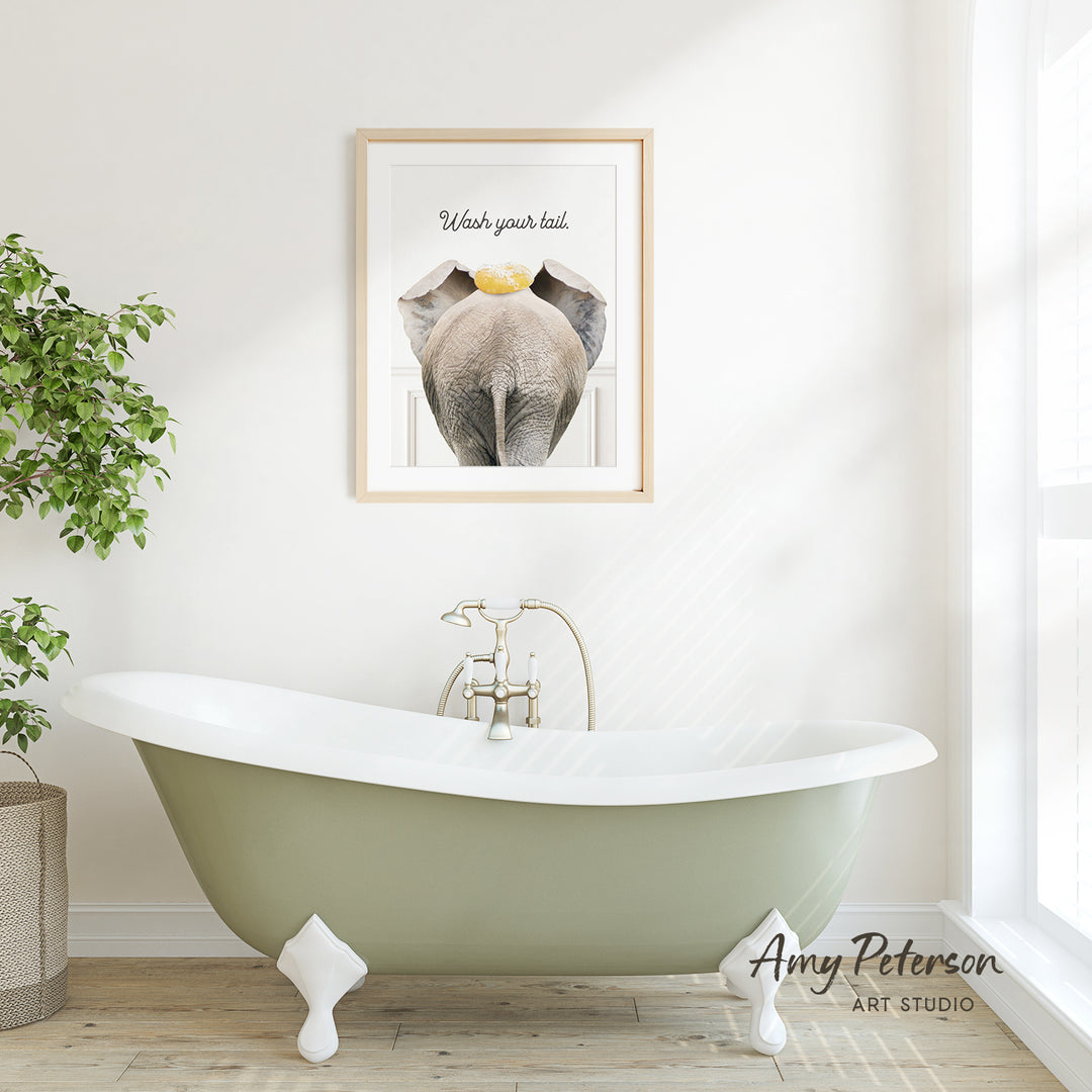 A framed poster of an elephant with the text "Wash your tail" is displayed above a green claw-foot bathtub in a bright, white bathroom.