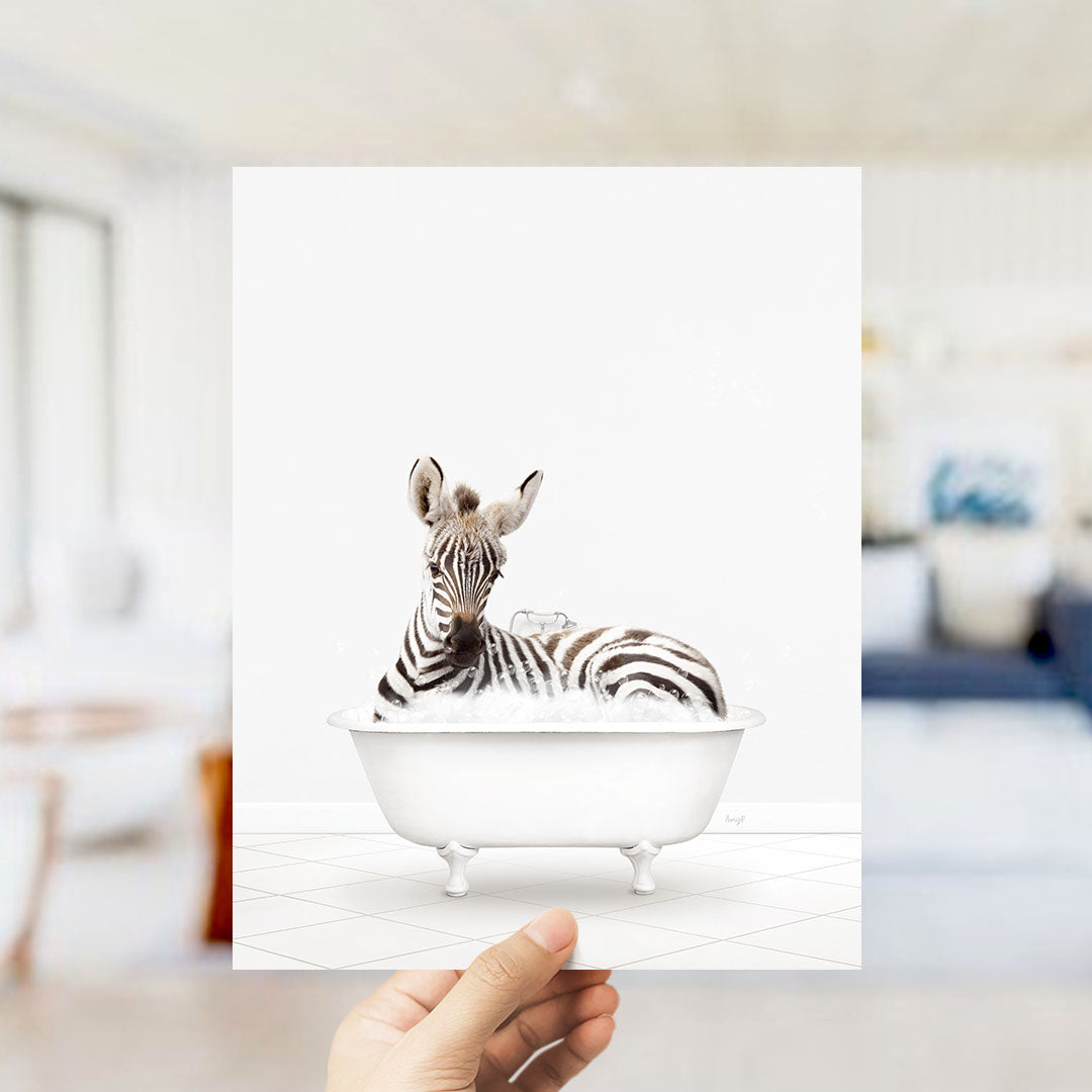 A hand holds up a framed image of a zebra sitting in a bathtub, surrounded by a blurred background.