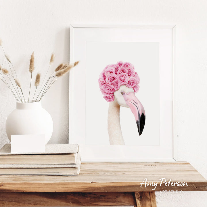 a framed picture of a flamingo with pink flowers on its head, placed on a wooden shelf next to a white vase with dried flowers.