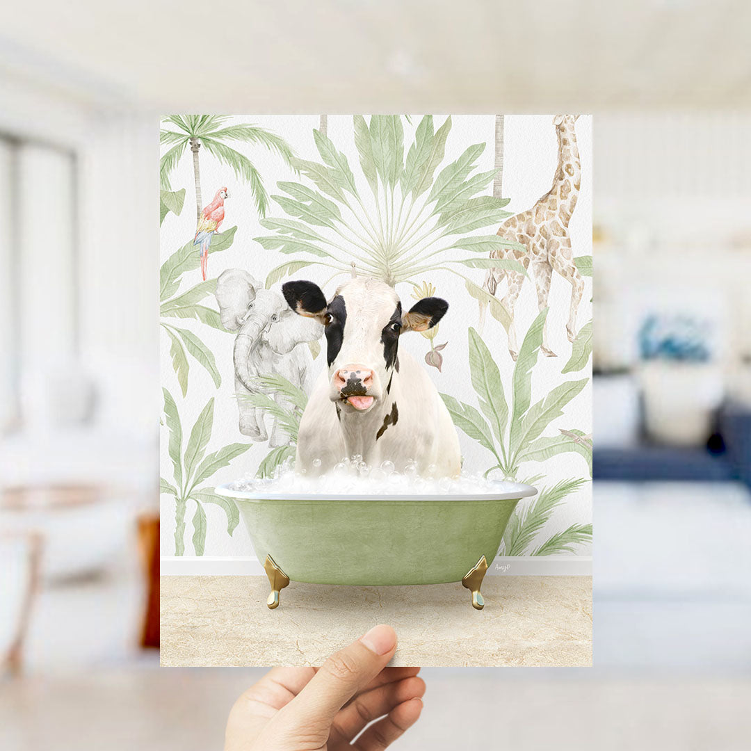 A hand holds up a framed print featuring a whimsical scene of a cow sitting in a bathtub surrounded by tropical foliage, including a giraffe and palm trees.