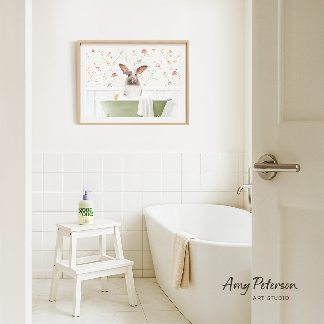 A framed image of a rabbit sitting in a bathtub is hanging on a white tiled wall above a white bathtub in a bathroom.