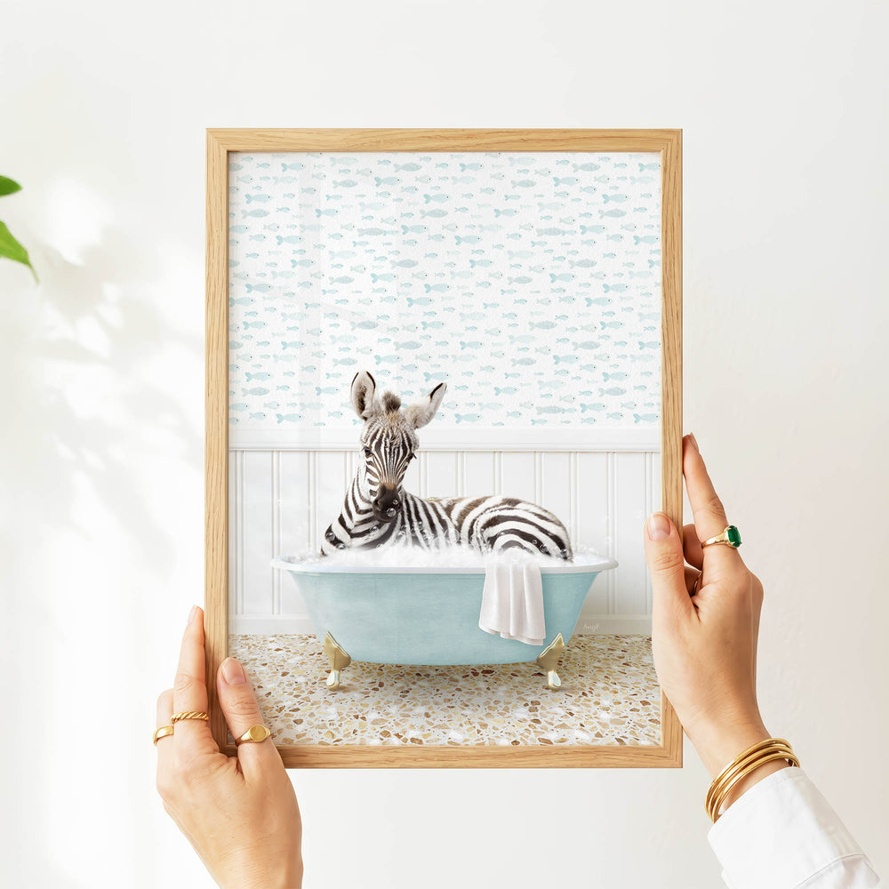 A person's hands are holding a framed picture of a zebra sitting in a bathtub filled with water, surrounded by a pattern of fish.