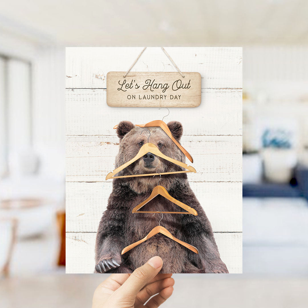 A hand is holding up a sign that says "Let's Hang Out on Laundry Day" with a bear sitting on a pile of clothes and hanging clothes on it.