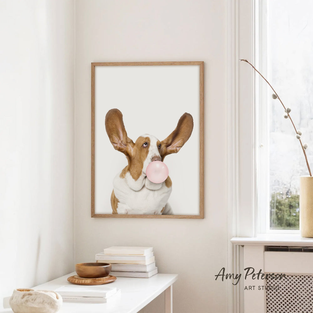 A framed image of a dog blowing a bubble gum bubble is hanging on a white wall in a room with a white table and a window.