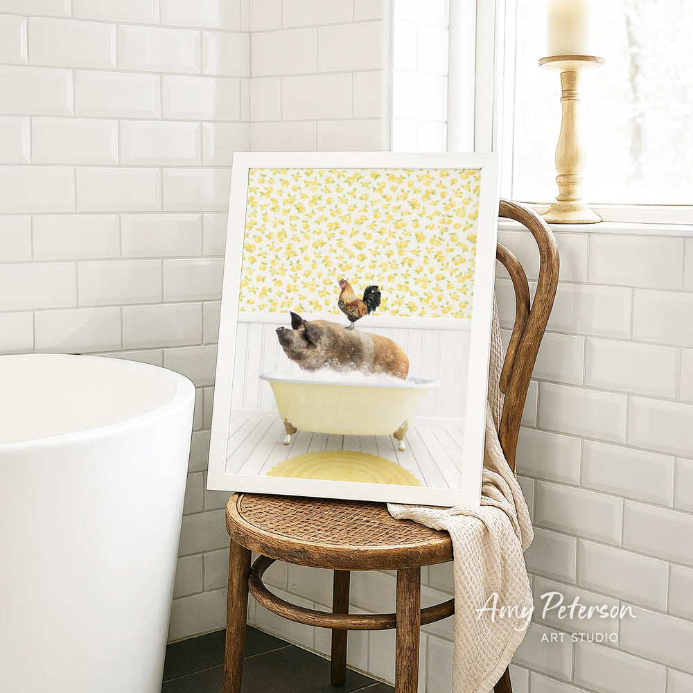 A framed image of a cat sitting in a bathtub, with a rooster perched on its head, is displayed on a wooden chair in a bathroom setting.