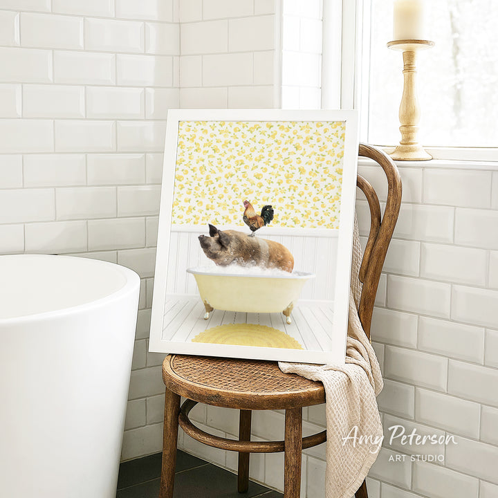 A framed image of a cat sitting in a bathtub, with a rooster perched on its head, is displayed on a wooden chair in a bathroom setting.