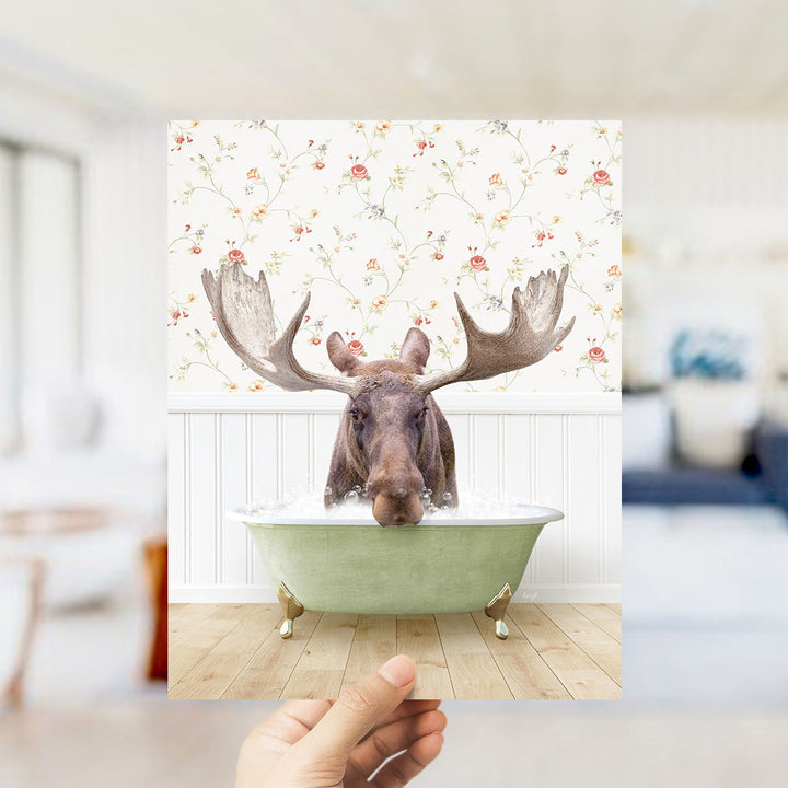 A moose with large antlers is sitting in a green bathtub filled with water, surrounded by a floral wallpaper.