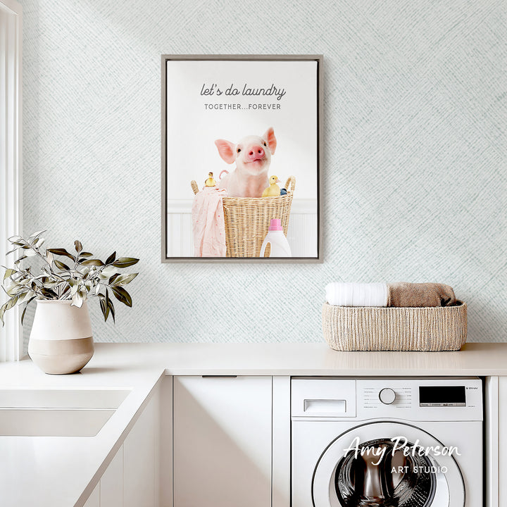 a framed poster of a pig sitting in a laundry basket, with the text "let's do laundry together forever" above it. The poster is hanging on a wall above a white sink and a white washing machine.