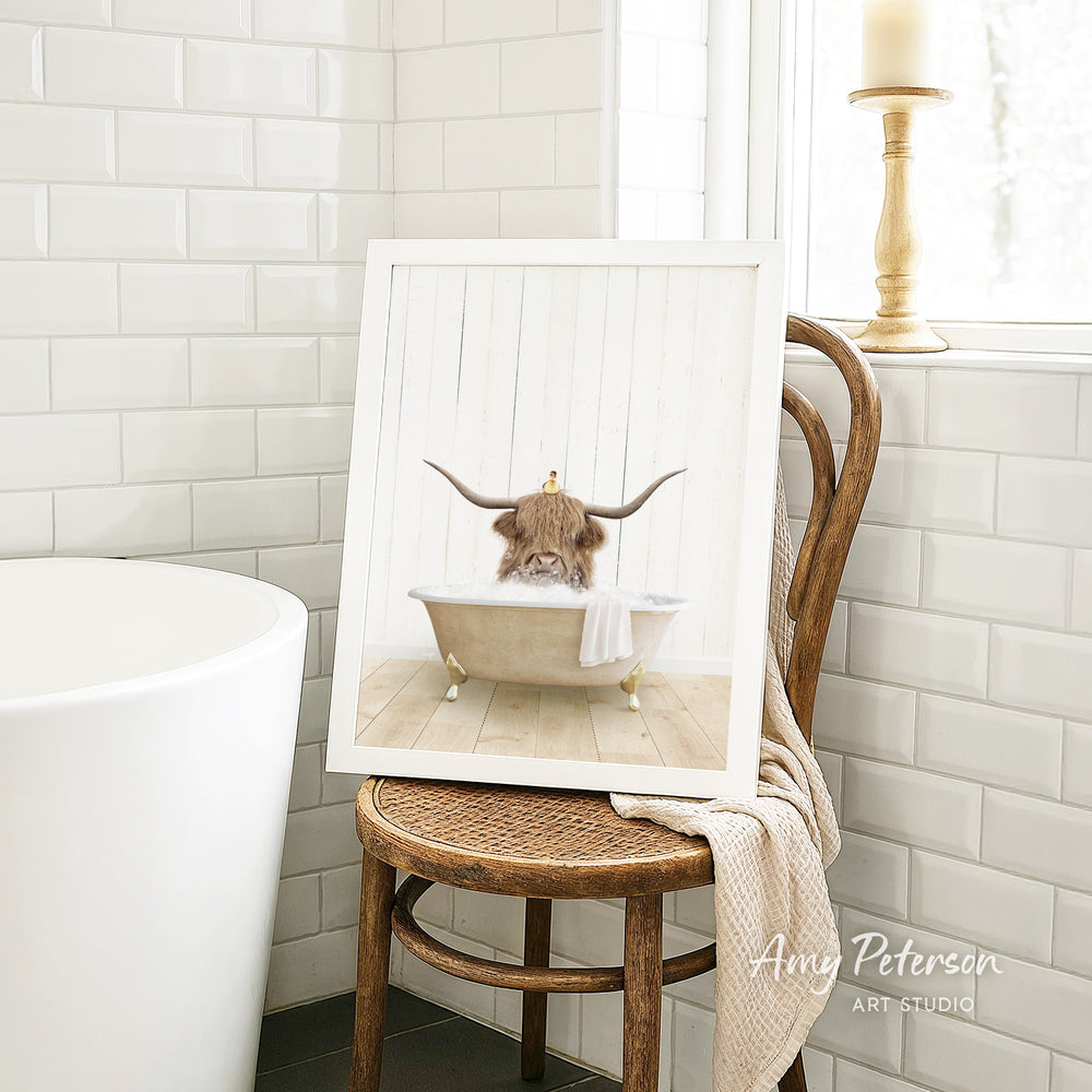 A framed image of a cow with horns sitting in a bathtub, surrounded by a wooden chair and a towel, is displayed in a bathroom setting.