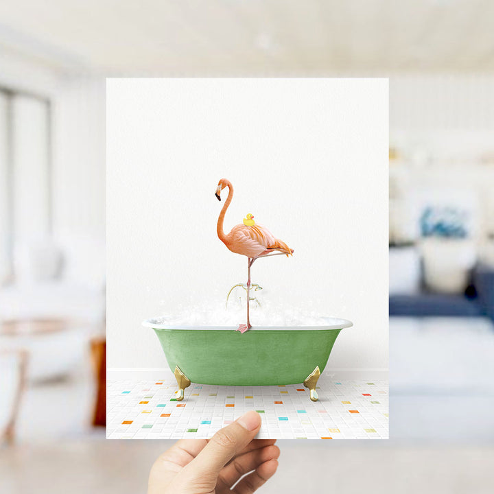 A hand holds up a framed image of a pink flamingo standing on a green bathtub filled with bubbles, with a yellow duck sitting on its back.