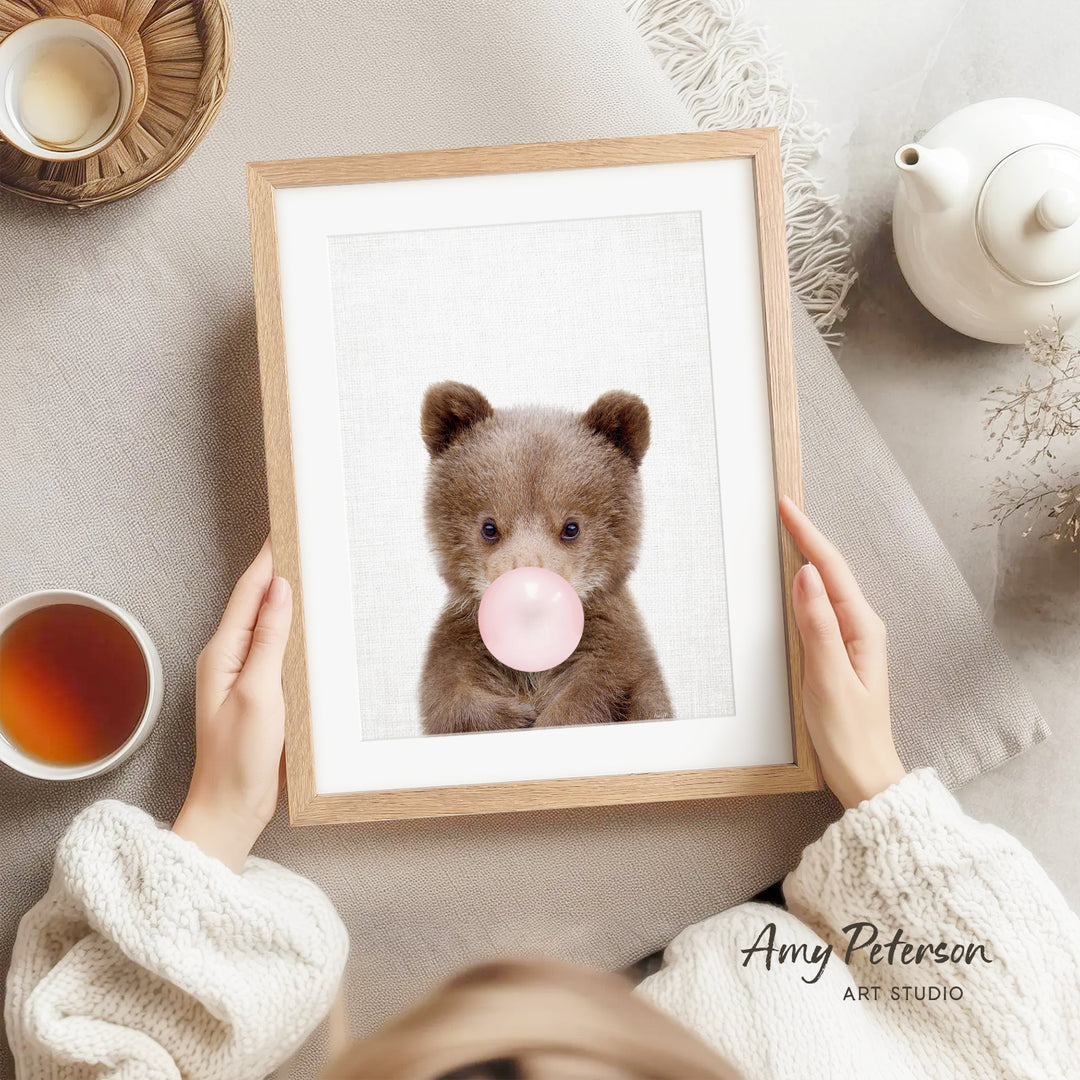 A person is holding a framed picture of a brown bear blowing a bubble with a pink bubble gum bubble.