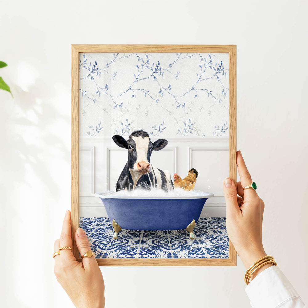 A framed image of a cow sitting in a blue bathtub, with a chicken nearby, is being held up by a person's hands.