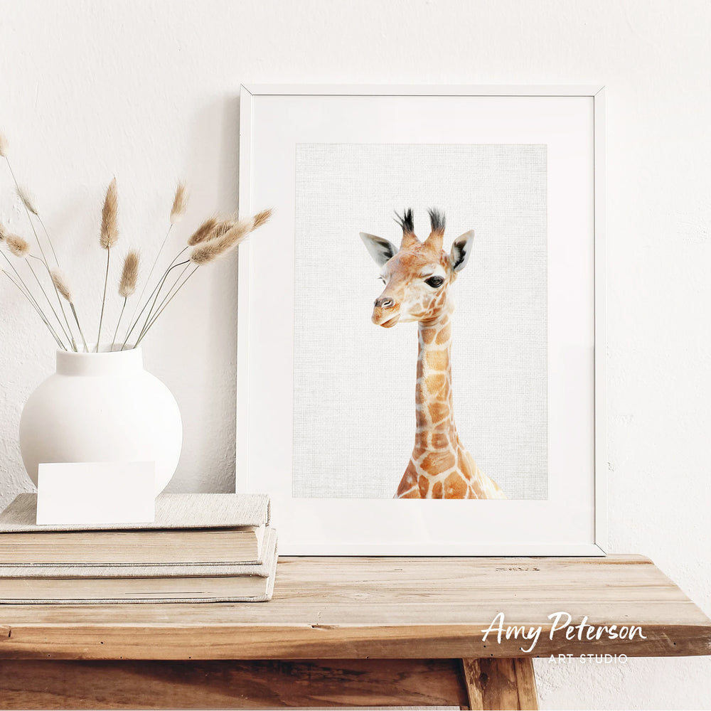 a giraffe in a white frame, placed on a wooden shelf next to a stack of books and a white vase with dried flowers.