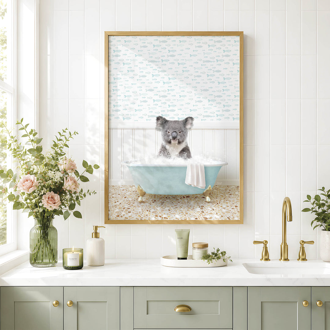 A framed image of a koala bear sitting in a bathtub, surrounded by a floral arrangement and a potted plant, is displayed on a white tiled wall above a bathroom sink.