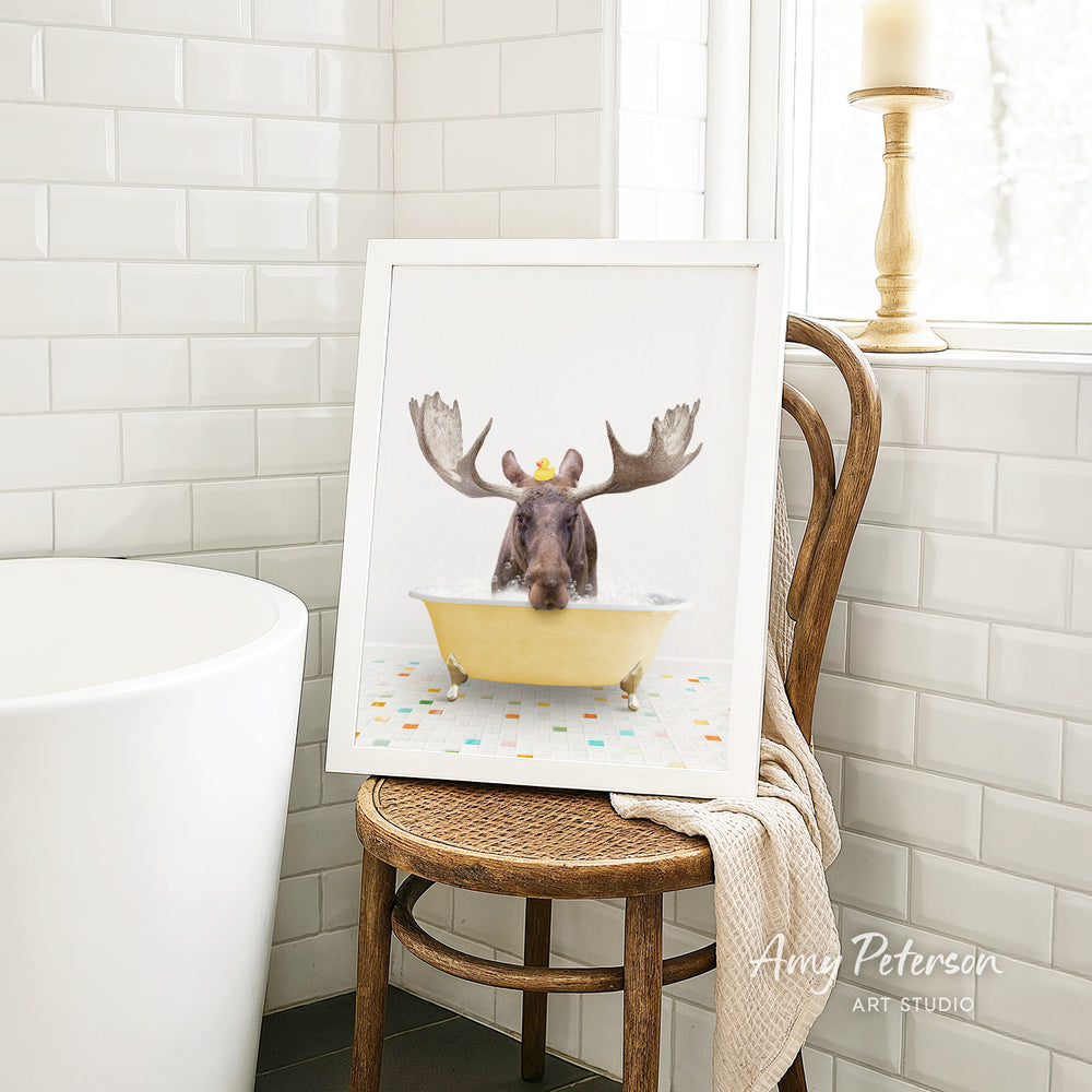 A framed image of a moose sitting in a bathtub, with its antlers raised, is displayed on a wooden chair in a bathroom setting.